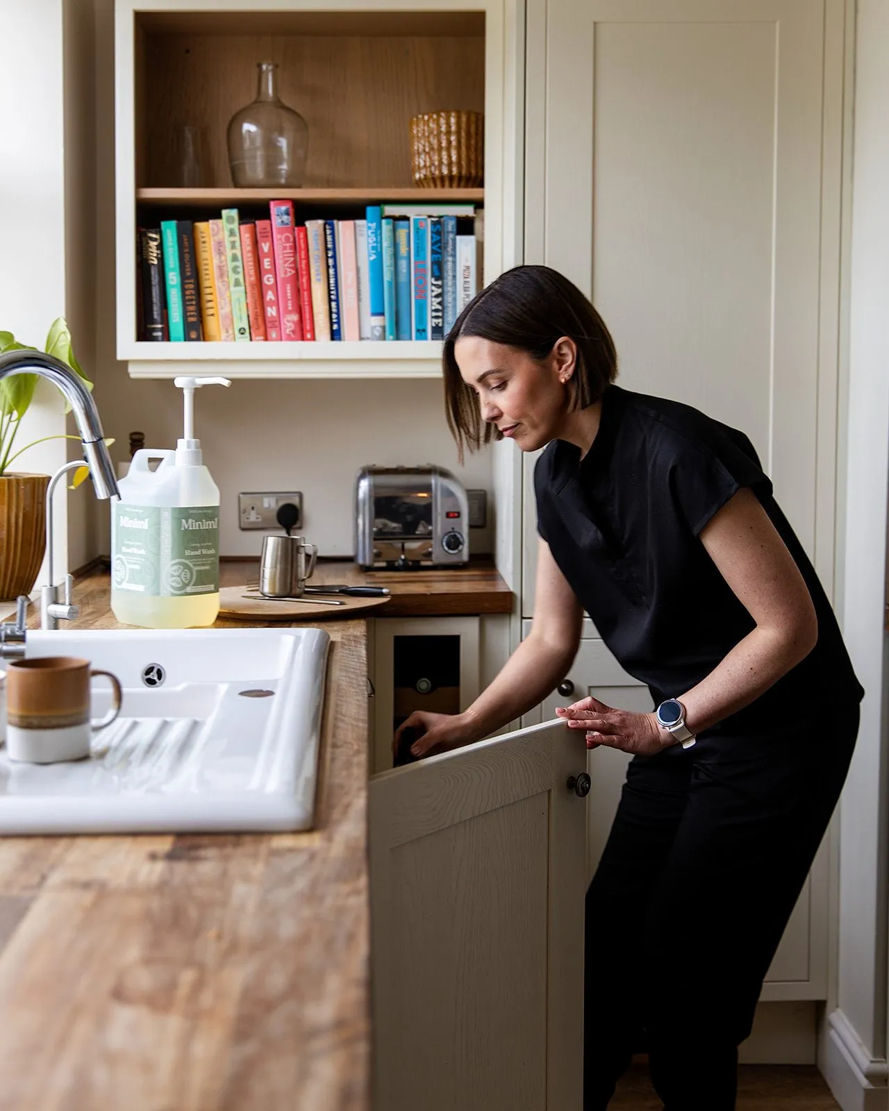 Grace closing a cupboard door in a bright kitchen with a butler sink.